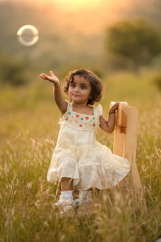 A toddler in a cream dress reaching up in a sunlit field